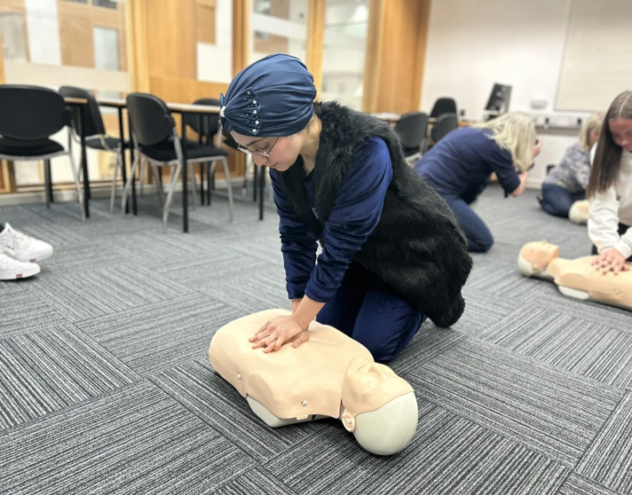 A woman wearing a blue headscarf and glasses practices CPR on a mannequin during a first aid training session in a classroom setting. Other participants are visible in the background. A woman wearing a blue headscarf and glasses practices CPR on a mannequin during a first aid training session in a classroom setting. Other participants are visible in the background.
